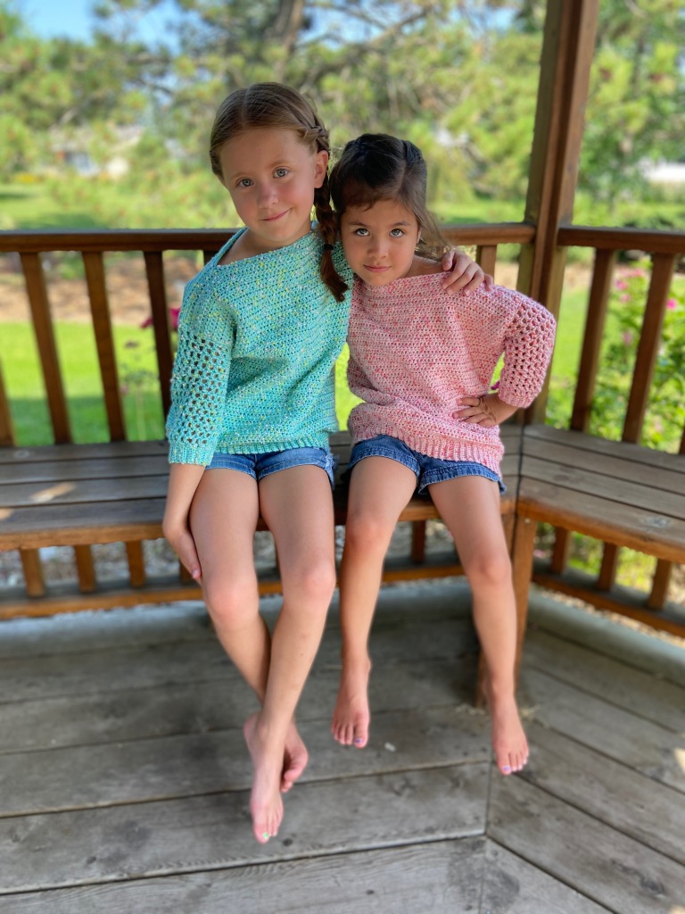 two girls wearing the breezy days pullovers in pink and seafoam sit o the bench of a wooden gazebo with gardens behind them, looking at the camera with an arm around each other